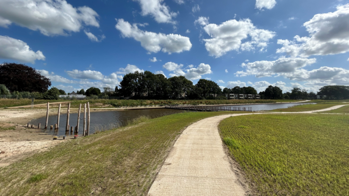 Een foto van gebied Vechtrijk Gramsbergen. De foto is gemaakt vanaf een looppad dat zich golvend door het landschap een weg naar rechts baant. Links van het pad en in de foto zie je een uitmonding van de Vecht, waarbij het uiteinde uitkomt bij een strand. Dit strand is verrijkt met klimpalen en andere speelelementen voor kinderen. Halverwege de uitmonding is een loopbrug die leidt naar de andere kant op het droge. Boven het land zien we een helderblauw lucht, vergezeld door enkele wolken.