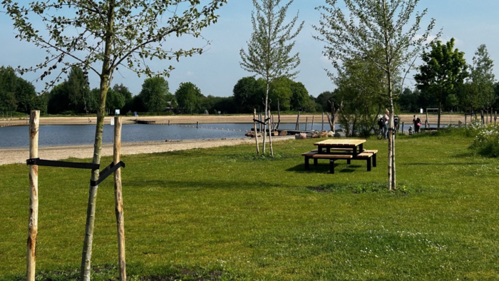 Een foto van de Kotermeerstal. Je ziet enkele jonge bomen op mooi groen gras. Iets verderop in beeld zie je een picknickbank. Helemaal achterin zijn enkele mensen te zien. Links in de foto zie je het strand en water.